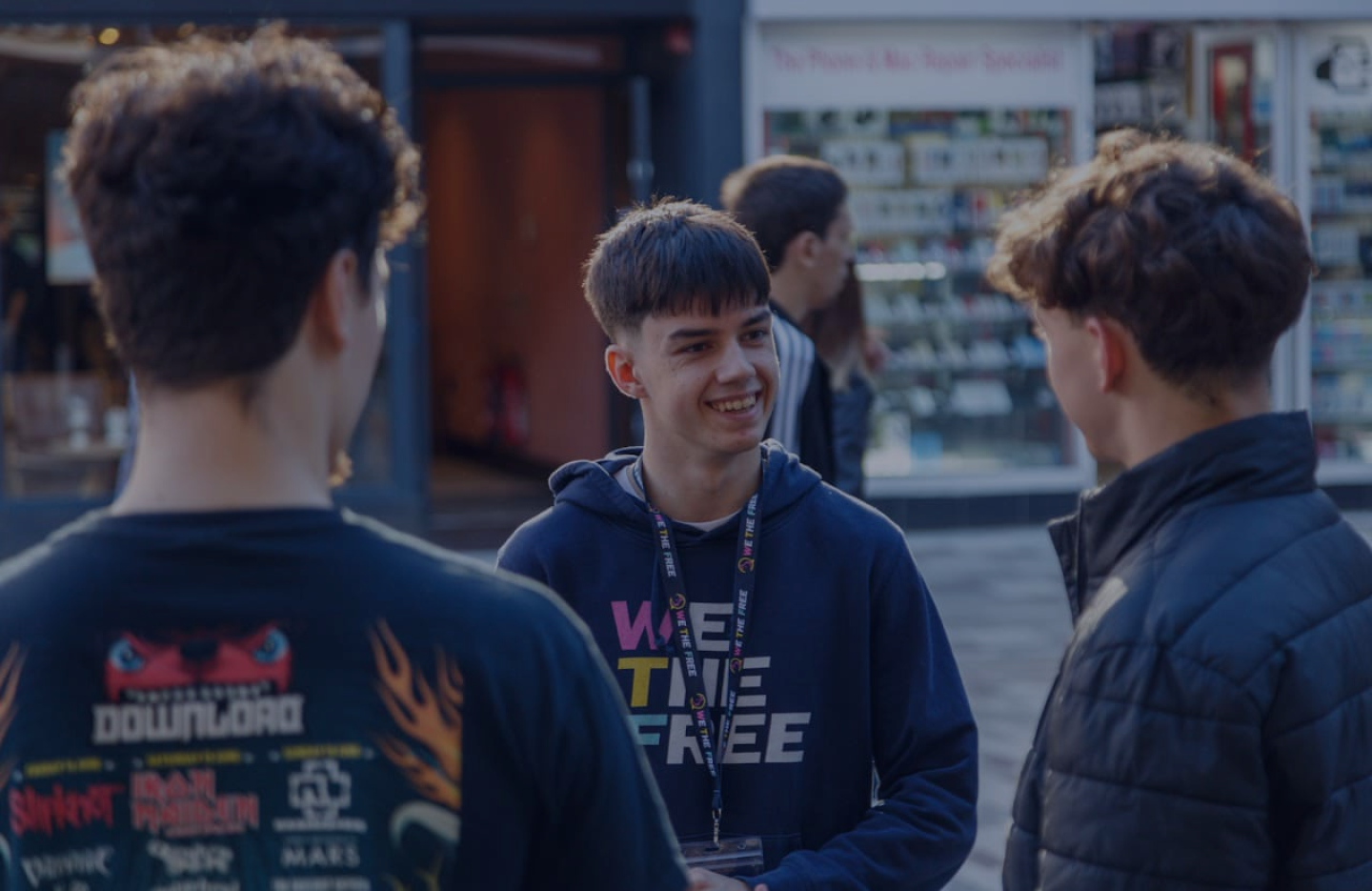 A We The Free volunteer in a branded hoodie and lanyard, smiling mid-conversation with two young people on a shopping street.