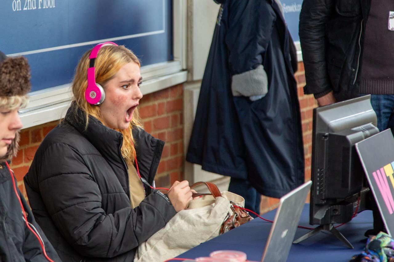A young woman in pink headphones watching a film at a WTF stall, mouth open in shock