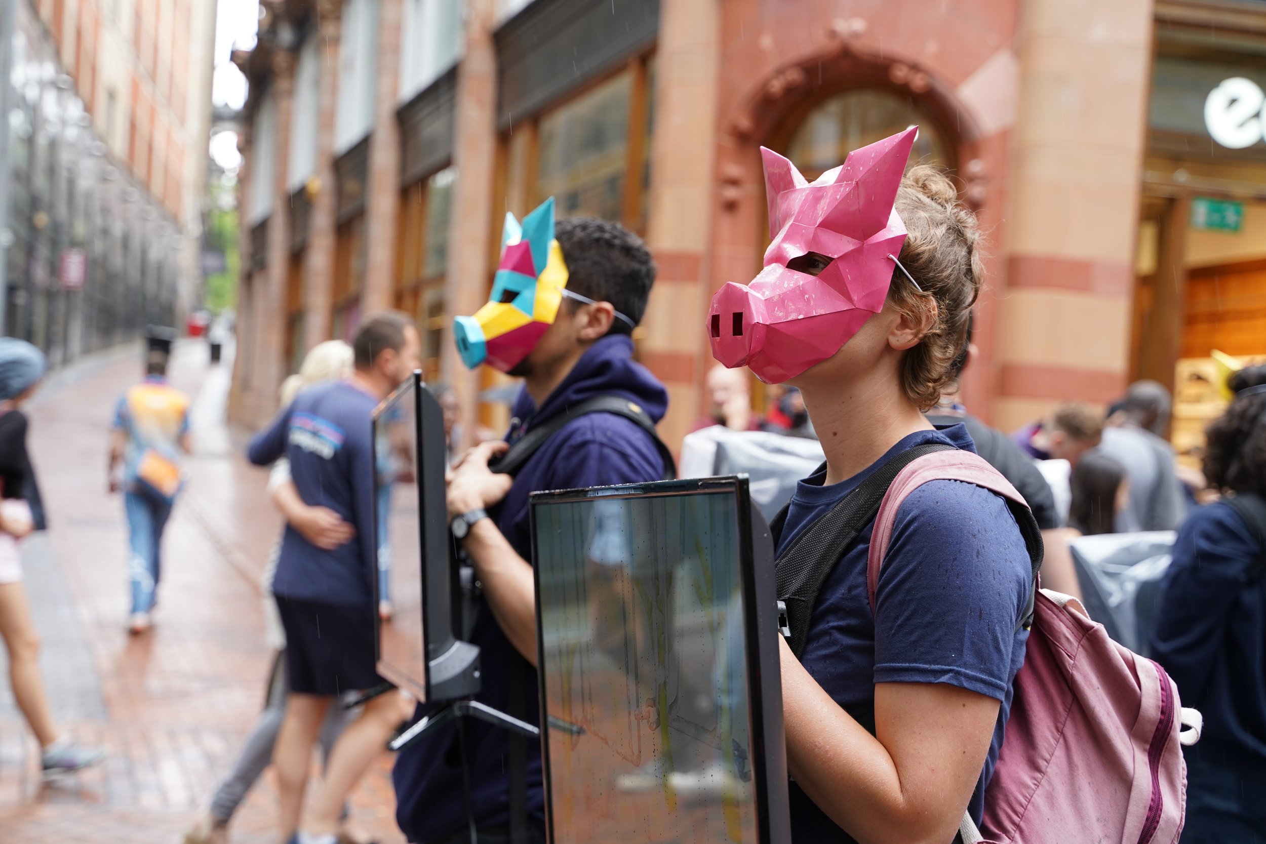 Two activists in geometric pig masks holding screens in a rainy shopping street