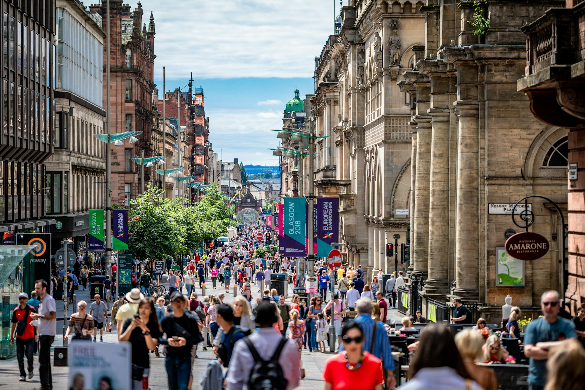 A busy pedestrian shopping street in Glasgow city centre on a sunny day, crowded with people between sandstone buildings